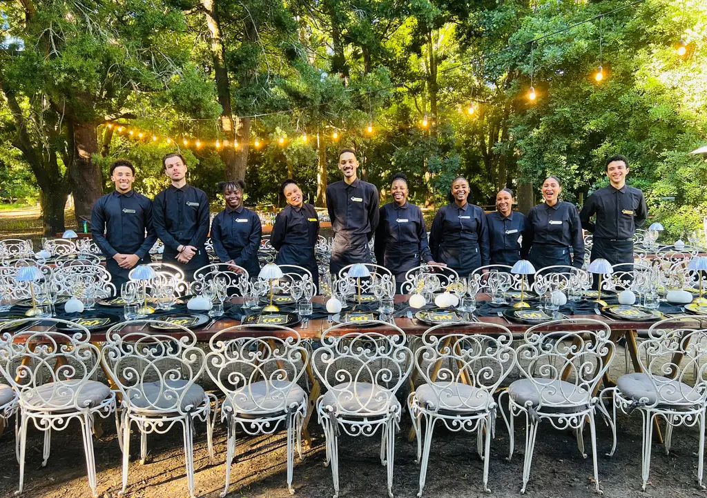 The Greyt-Waitstaff team in black uniforms lined up behind a long dressed table under string lights in a forest clearing