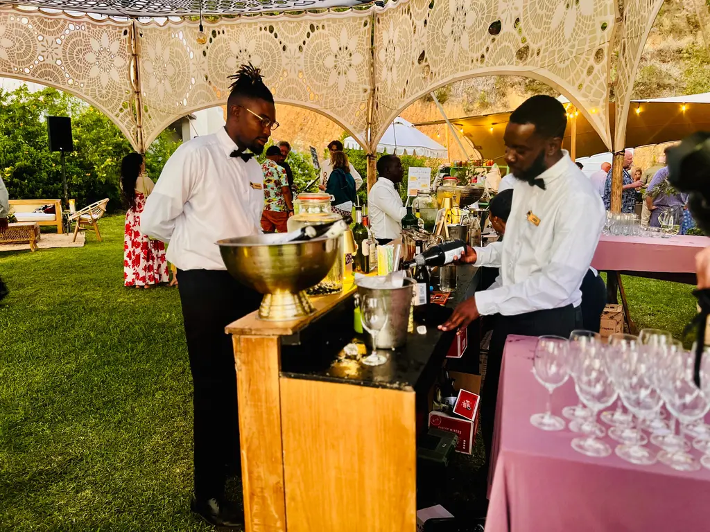 Two bartenders in bow ties pouring drinks at a wooden bar under a lace tent