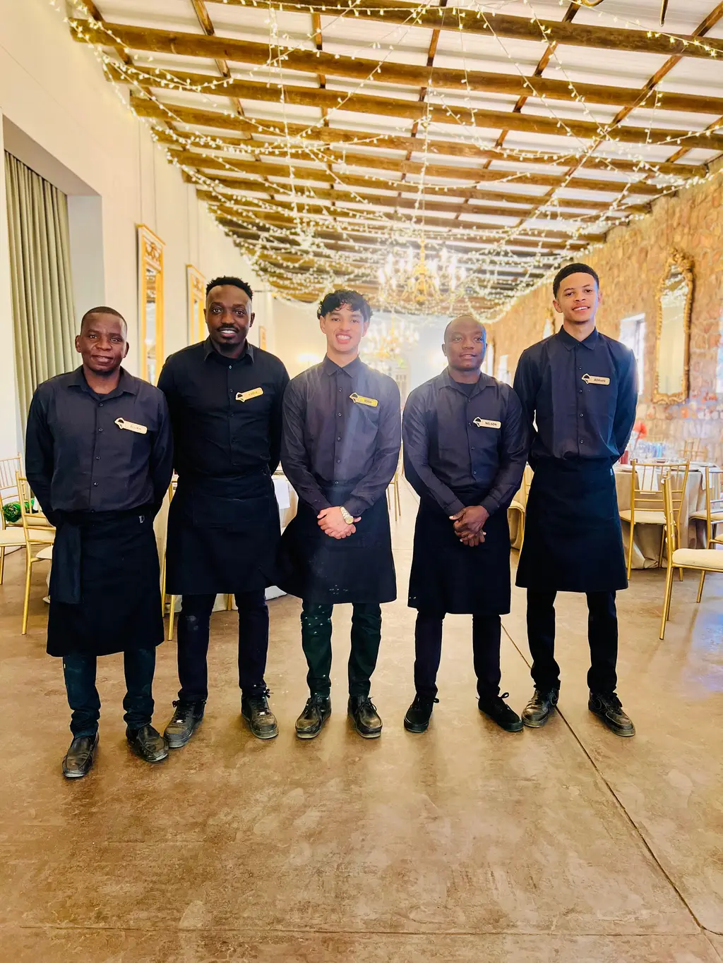 Five waiters in black standing in a hall with fairy-lit beams and chandeliers