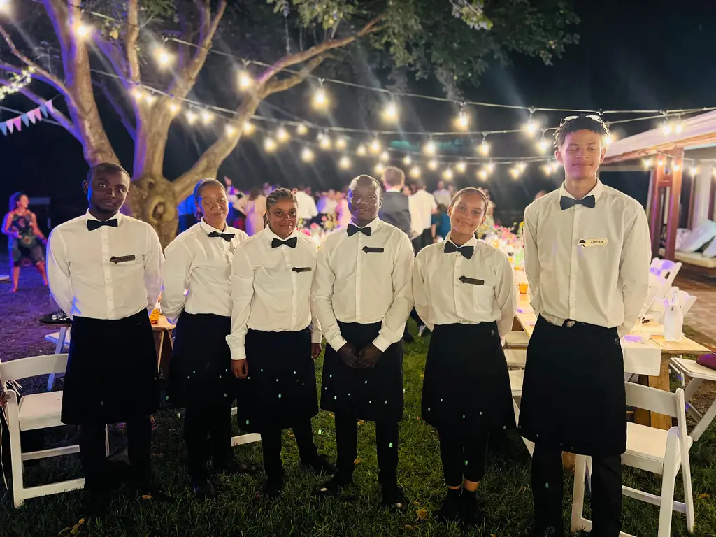 Six bartenders and waiters in white shirts and bow ties standing under festoon lights at a night-time party
